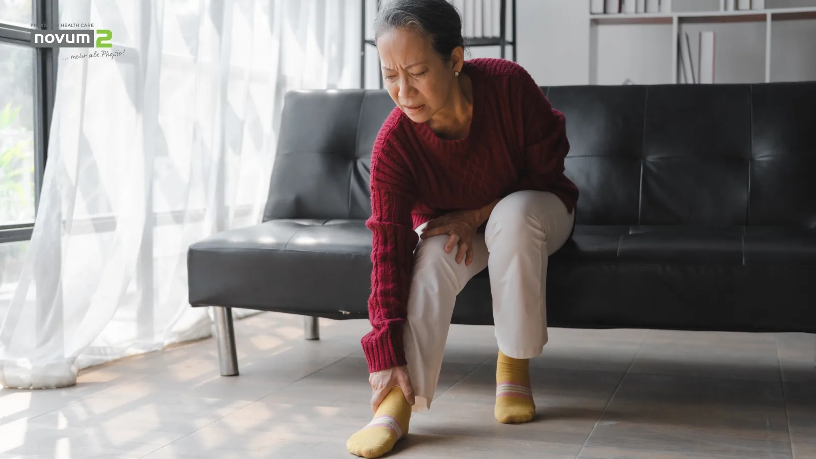 Elderly woman holding her aching foot while sitting on a sofa at the Novum2 physiotherapy practice